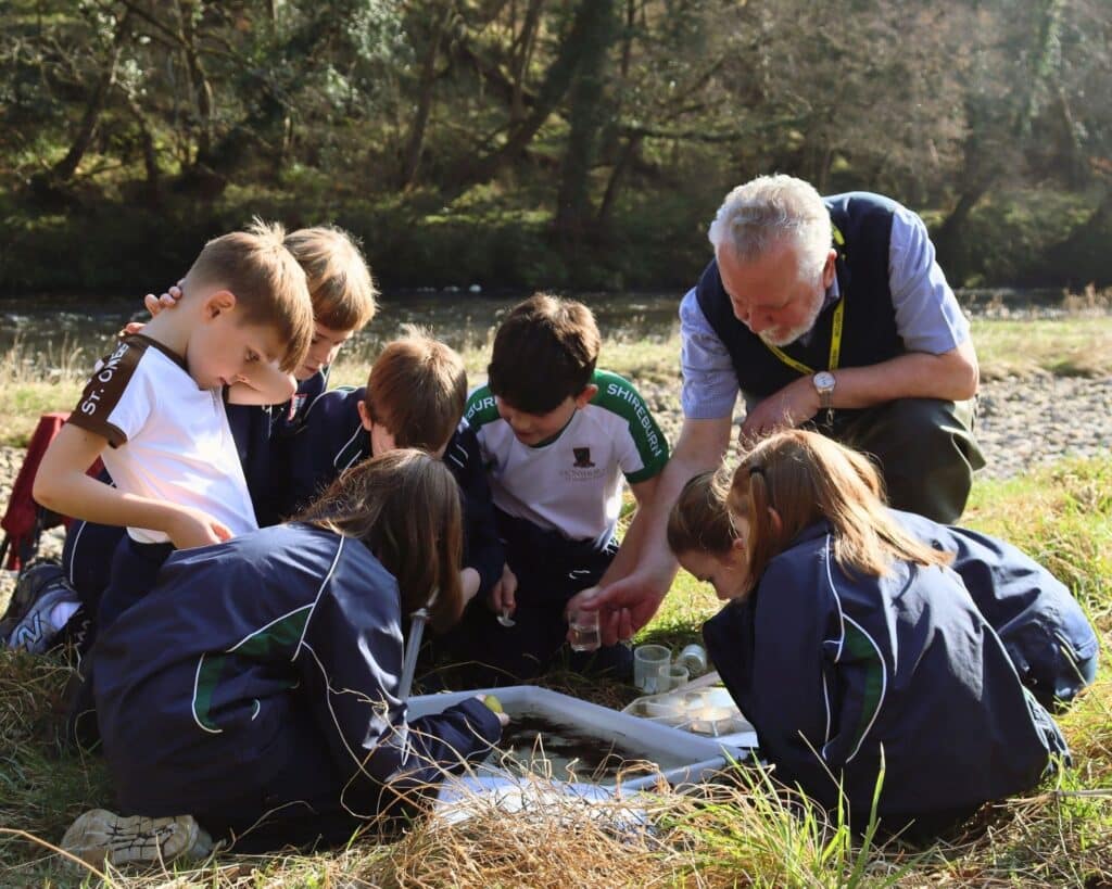 An education volunteer kneels beside seven children- together they are examining invertebrates in a tray next to the River Hodder. Community engagement and education is a huge part of Hodder Together for Salmon
