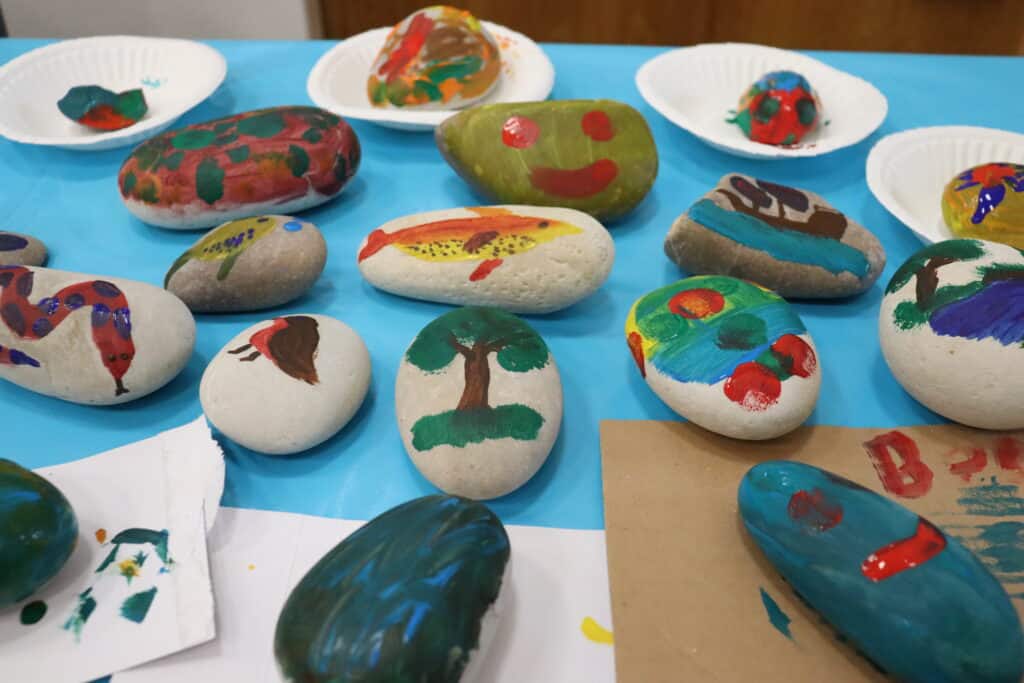 Painted pebbles lie drying on a table. Each pebble has a nature themed painting on it including trees, rivers, fish, and birds.